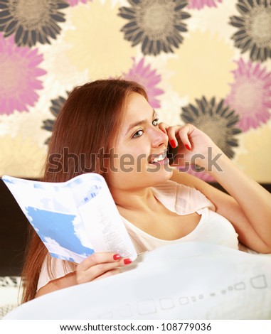 A teenager girl lying on a bed reading a book and talking on a mobile phone at the same time