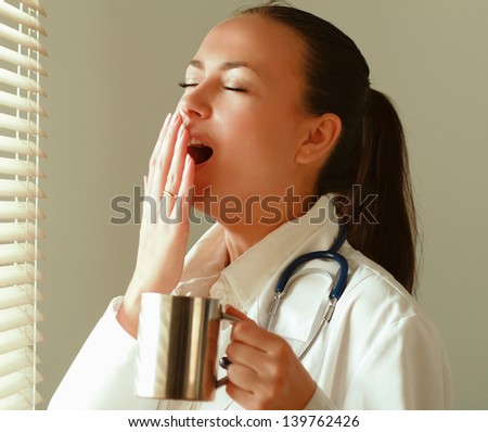 The  yawning woman is standing in the office near window and holding cup