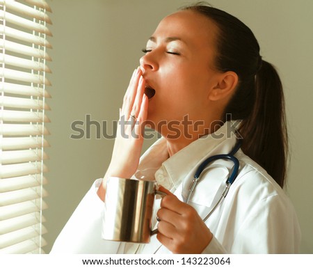 The  yawning woman is standing in the office near window and holding cup