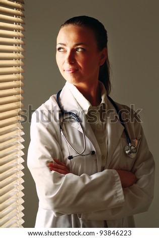 Woman doctor is standing near window with folded arms