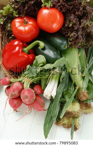Fresh corn, red leaf lettuce, tomatoes, cucumbers, red pepper, radishes and green onions sit in a basket on an aged table