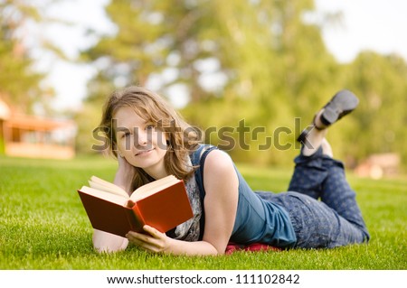 Charming girl lies on green grass and reads book.