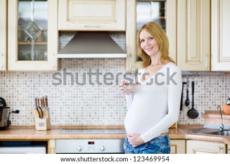 Attractive pregnant woman holding a glass of clean water while standing in the kitchen and looking at camera