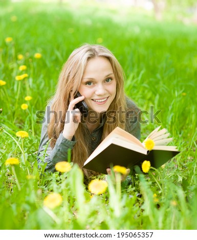 girl lying on grass with dandelions reading a book and talking on the phone