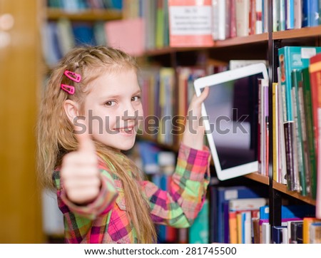 Happy teen girl pulls a tablet pc off the shelves in the library and showing thumb up