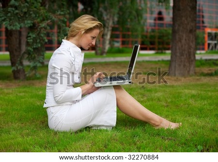 Young women sits on a grass with laptop, looks at it and type