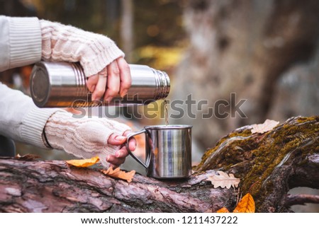 Woman with knitted glove is pouring a hot drink into mug from thermos. Refreshment during hiking.