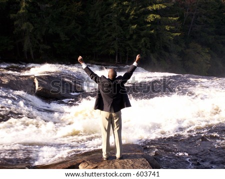 business man in a suit with cell phone standing in front of a rushing river with fists raised in a triumphant gesture
