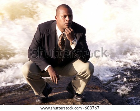 business man in a suit squatting down thinking in front of a rushing river
