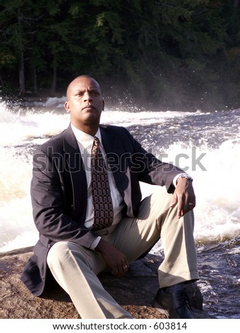 business man in a suit sitting on a rock in front of a rushing river