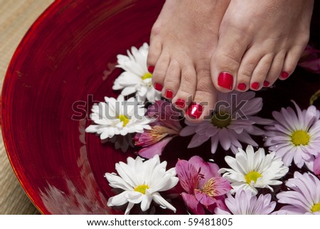 A women with red nail polish on her toes holds her feet above a bowl of water and flowers.