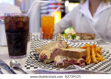 A Reuben sandwich with sweet-potato fries rests on a table across from an out-of-focus woman.