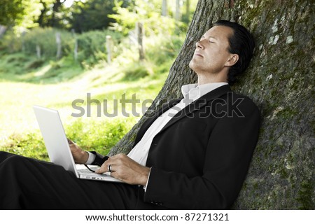 Businessman relaxing against a tree with his laptop