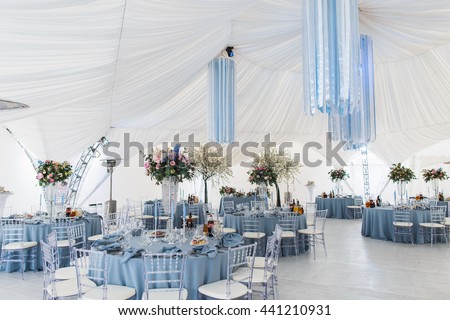 Round dinner tables covered with blue cloth stand in a white wedding pavilion