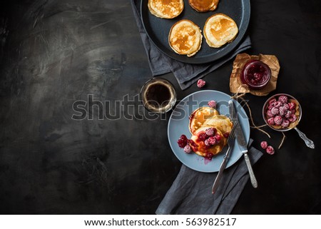 Pancakes with raspberry jam on a blue plate, frozen raspberries, a jar of raspberry jam, a cup of coffee, a frying pan with pancakes, fork and knife on dark metal table top view. Breakfast.