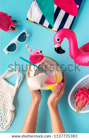Flat lay of summer vibes concept with colorful travel items, sunglasses, scarf, coconut water, pink dragon fruit, flamingo inflatable drink holder and net bag on a blue background.