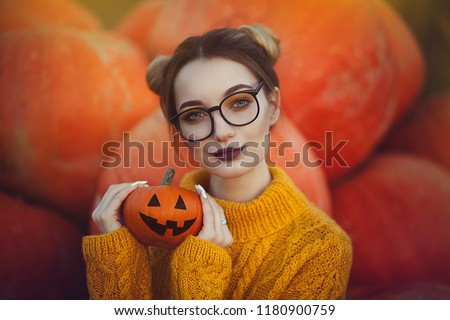 A cozy autumn photo of a girl with pumpkins in a yellow sweater and glasses. Stylish young woman in autumn decorations, in hands a Halloween pumpkin.