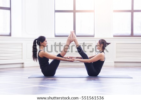 Two attractive sport girls work out nauka asana boat yoga pose on black mat in fitness class. Group of young women stretching in gym with windows