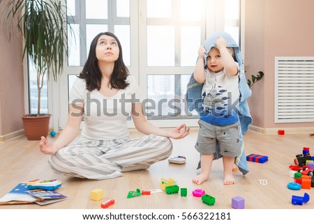 Portrait of young tired yoga mom spending time with her little baby boy, relaxing while her kid son playing games, throw toys and arrange a mess at home