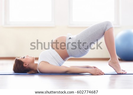 Portrait of a beautiful young pregnant woman holding her stomach while sitting near an exercise ball in the sport center. Working out, yoga and fitness, pregnancy concept.