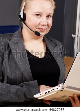 Woman at a helpdesk with white laptop and headset, dressed in a business suit, serious while looking at the camera