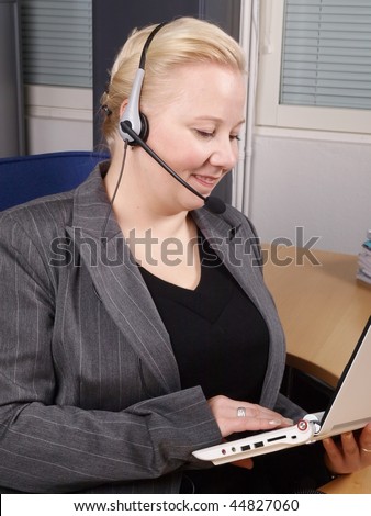 Woman at a helpdesk with white laptop and headset, dressed in a business suit, smiling while watching the PC