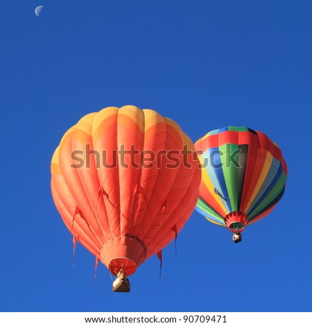 two hot air balloons soar into clear blue sky, with moon