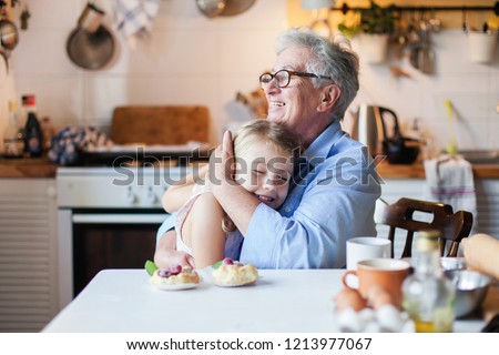 Happy grandmother is hugging granddaughter in cozy home kitchen. Family is cooking together. Senior woman and cute little child girl are smiling. Kid is enjoying kindness, warm hands, care, support.