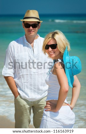 Happy young couple in sunglasses and white dress on a tropical beach with blue sea on background. Selective focus on the girl.