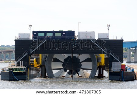 Giant tidal turbine docked in a harbor. It is held in a ship designed to hold and transport it. Sky is overcast. Walkways on ship give sense of scale. Identifying marks removed. Room for text.