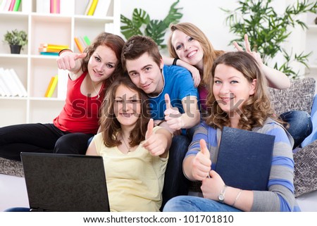 Group of five young people with thumbs up sitting on white sofa, working on laptop computer indoors.