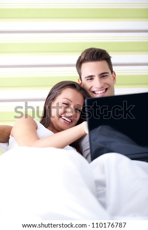 Happy couple with laptop in bed watching fun movie on computer in bed before sleeping. Smiling young interracial couple in bed at home.