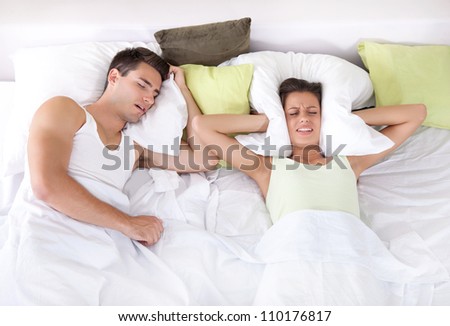 Couple in bed; man snoring and woman can not sleep; covering ears with pillow for snore noise.