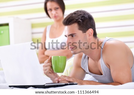 Man at computer, women upset and angry looking at man. Young modern interracial couple in bed.