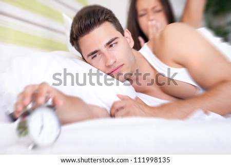 Young snoozing couple in bed waking up to alarm clock.