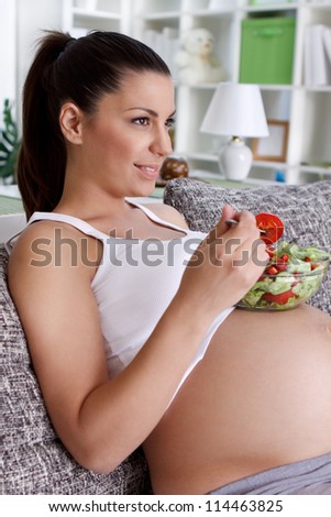 Beautiful pregnant woman eating salad and relaxing in the living room