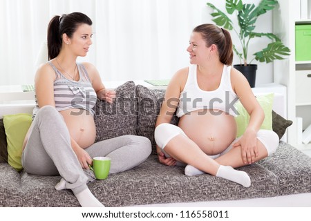 Two pregnant female friends sitting on sofa at home and sharing experiences