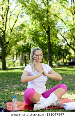 Beautiful blond woman practicing yoga in the park on a green grass, sitting on mats with her legs crossed.