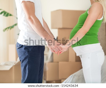 Cute pregnant wife holding hands of  her husband standing front of boxes during moving in new home