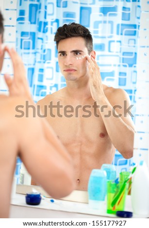young man applying moisturizer on his face front of mirror in bathroom