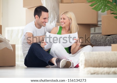 Attractive couple sitting on home floor looking at house plans and smiling at each other.