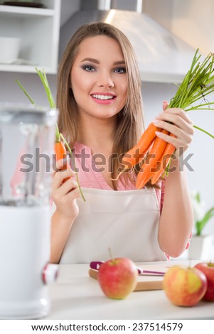 Young cheerful woman in kitchen holding carrots, ready to making fruits smoothies with blender