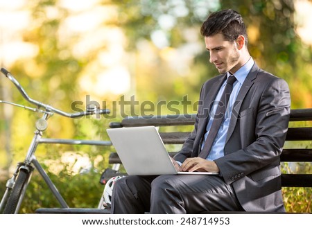 Businessman with bicycle working on laptop on bench in park