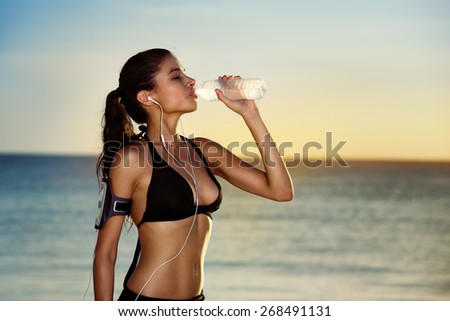 Fitness beautiful woman drinking water and sweating after exercising on summer hot day in beach. Female athlete after workout
