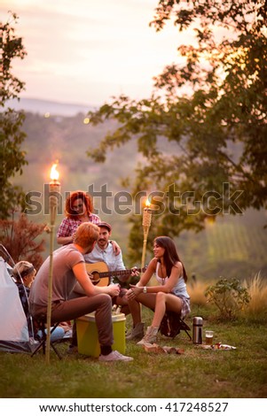 Guys and lassies talk and play guitar at twilight in campground in nature