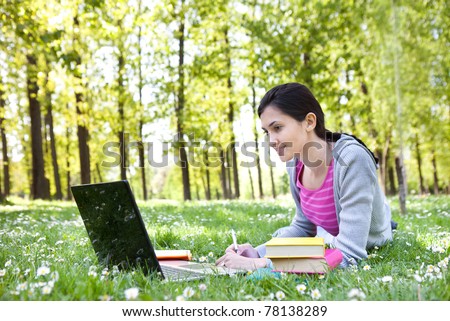 beautiful young student girl lying on grass with laptop and books, looking into the laptop and smiling