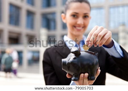 beautiful young woman putting a coin into a big pink piggy bank in front her office building (focus on the piggy bank)