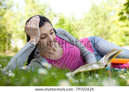 young woman with headset listening music and reading book in park, lying on grass