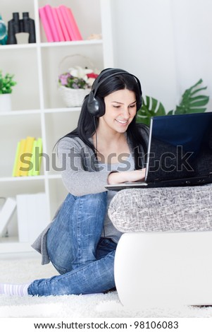 Pretty woman using laptop computer and headphones, typing, looking at screen, sitting on living room floor at home.