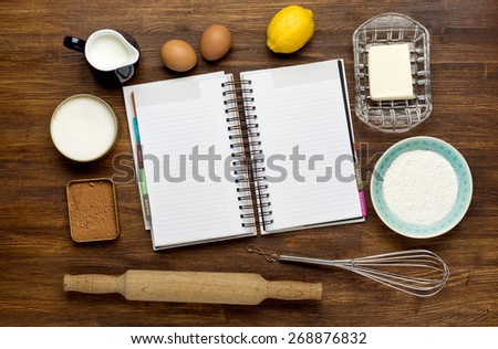 Rural vintage wooden kitchen table with blank recipe book and baking cake ingredients (chocolate, eggs, flour, milk, butter, sugar). Background layout with free recipe text space.